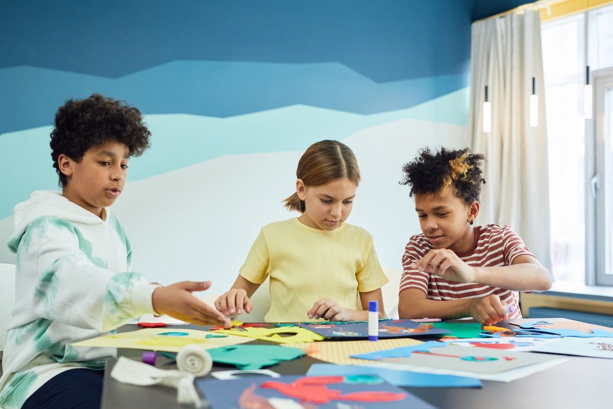 Three children making paper crafts together at a bright classroom table