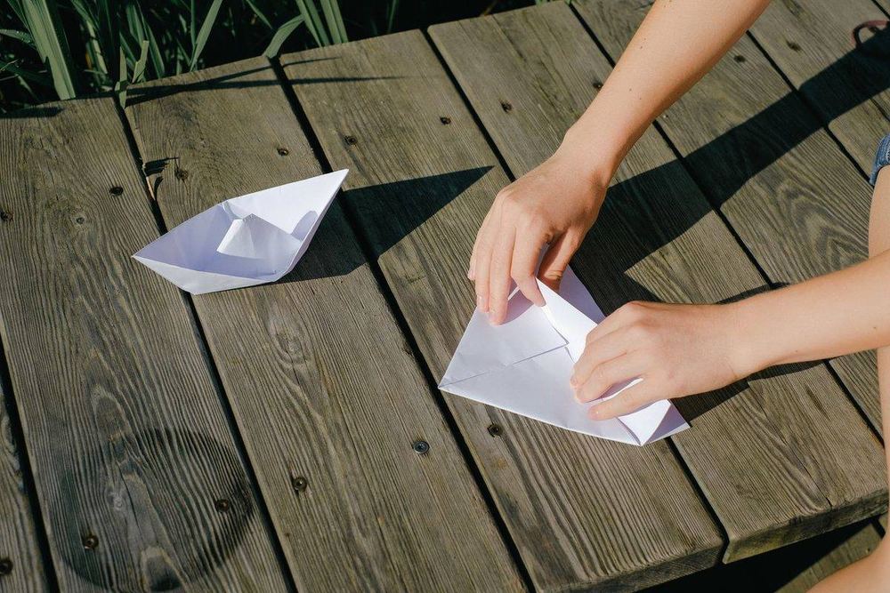 Hands folding a paper boat on weathered decking beside a finished one