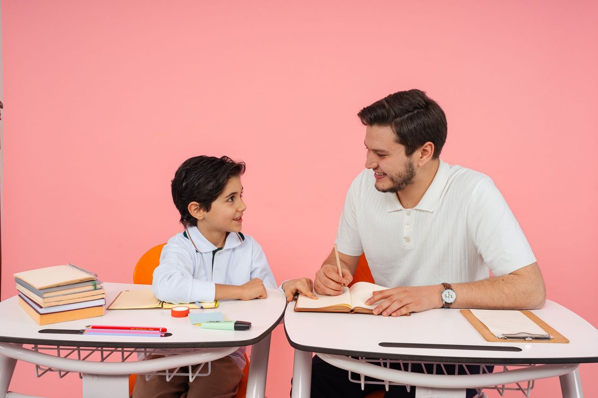 An adult sitting opposite a child helping with written work at a desk